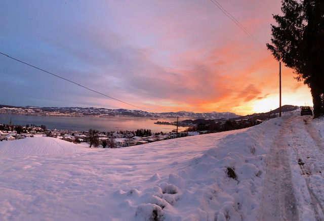Die Landschaft ist verschneit, im Hintergrund ist ein See und dahinter Berge zu sehen. Der Himmel wird von Rot-, Gelb- und Orangetönen dominiert. Es ist Sonnenaufgangszeit.
