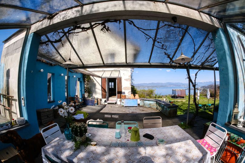 Das Bild ist mit einem Fischauge-Objektiv fotografiert. Es ist eine Terrasse zu sehen, im näheren Hintergrund das Haus, welches über die Terrasse erreicht werden kann. Weiter im Hintergrund ist ein See zu sehen. Die Terrasse ist bunt gestaltet.